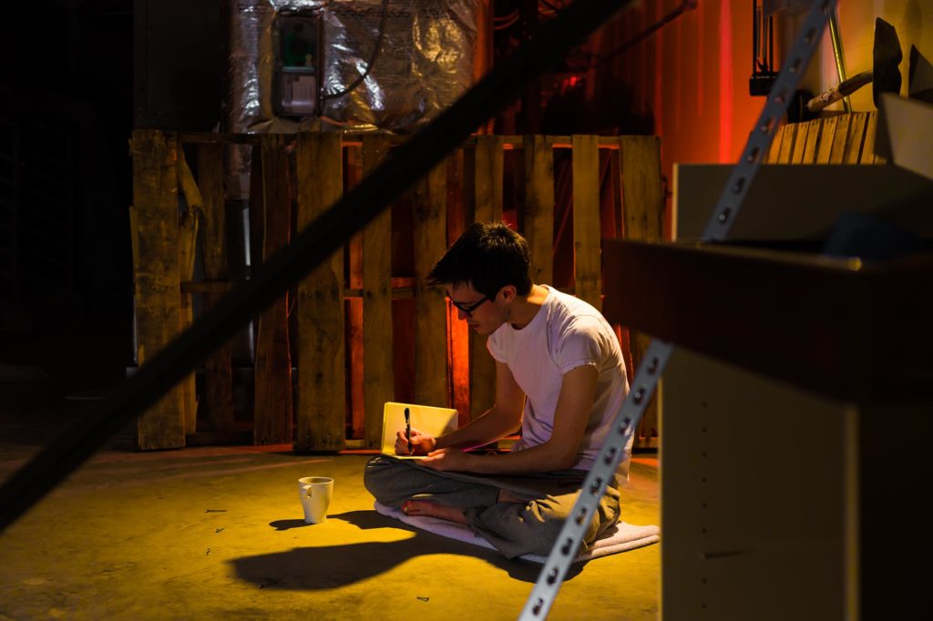 Picture of Nicholas Ryan in an industrial setting with red lighting writing in in a notebook with a cup of coffee next to him.